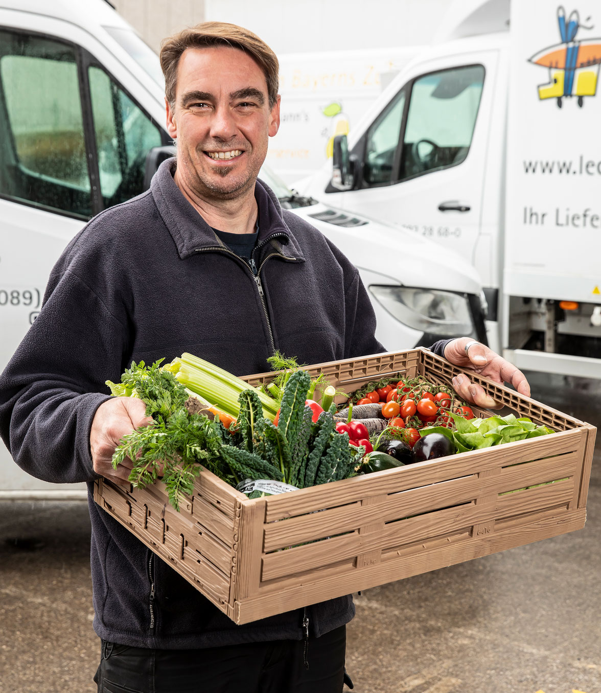 Ein Lieferant von Lunemann's® in dunkelblauer Arbeitskleidung hält eine Kiste mit frischem Gemüse, darunter Sellerie, Tomaten und Salat. Im Hintergrund stehen weiße Lieferwagen mit einem Logo und der Aufschrift "Ihr Lieferservice".