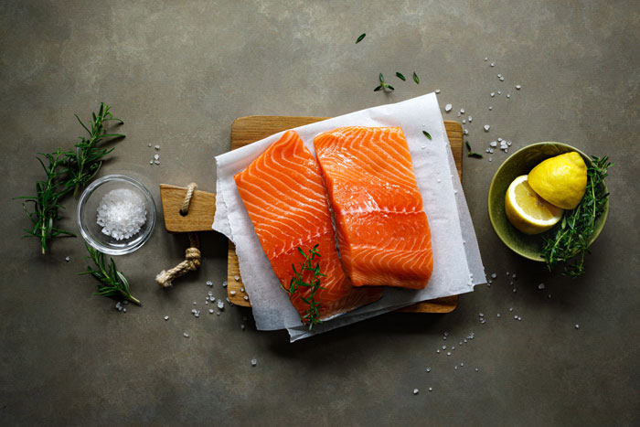 Geräucherter Lachs auf einem Teller, serviert mit frischen Kräutern und einer Zitronenscheibe, auf grauem Untergrund.