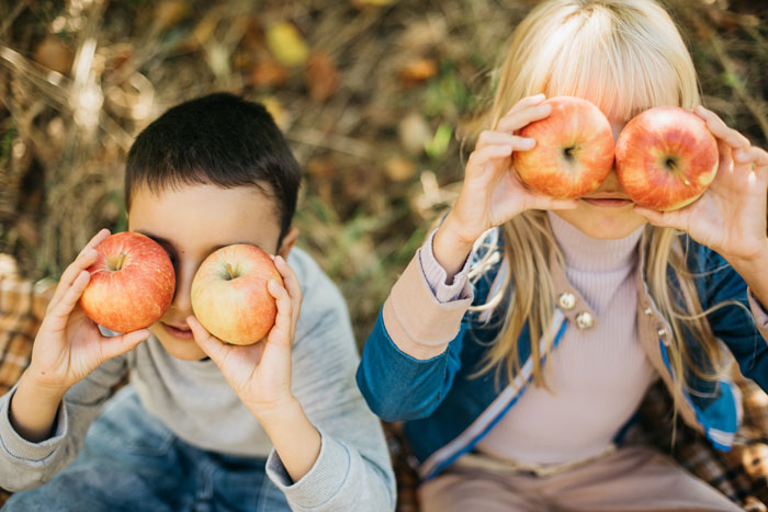 Zwei Kinder halten jeweils einen Apfel vor ihr Gesicht und lächeln spielerisch in einem herbstlichen Garten.