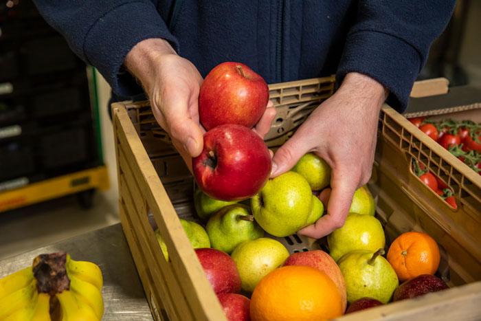 Hände halten frisches Obst, darunter rote Äpfel, grüne Äpfel und Orangen, in einer Holzkiste.